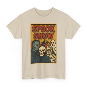 May include: A beige t-shirt with a retro-style graphic. The design features a Frankenstein's monster, a skull, a mummy, and a ghost, with the words "SPOOK SHOW" in a vintage font. The shirt is made of a soft material.