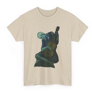 以下が含まれることがあります： ギターを弾く人物が描かれたベージュ色のTシャツ。人物は青と緑の色合いで描かれ、顔色は青白い。ギターは茶色と黄色。Tシャツはクルーネックです。