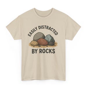 May include: Beige t-shirt with the text "EASILY DISTRACTED BY ROCKS" in black. The graphic features a collection of cartoon rocks in shades of gray, brown, and blue, resting on a patch of sand.