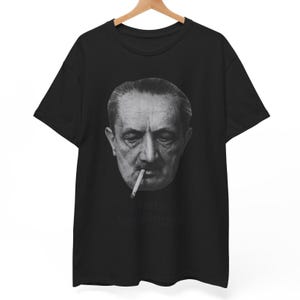 May include: Black t-shirt featuring a grayscale image of a man with a cigarette in his mouth. The image is a close-up of the man's face. The text "Martin Heidegger" is printed below the image in a stylized font.