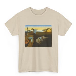 May include: A beige t-shirt featuring a surrealist painting by Salvador Dalí. The artwork depicts melting clocks, a barren landscape, and a coastal scene. The shirt is a classic crew neck style.
