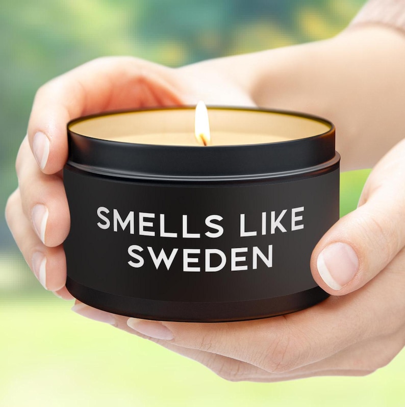 K&ouml;nnte beinhalten: Eine brennende schwarze Kerze in einer runden Dose mit dem Text "Smells Like Sweden" in Wei&szlig;. Die Kerze hat eine goldfarbene Innenseite und wird von zwei H&auml;nden gehalten. Der Hintergrund ist verschwommen mit Gr&uuml;n- und Gelbt&ouml;nen.