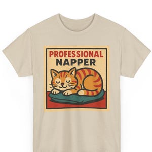 Könnte beinhalten: Beiges T-Shirt mit einer Grafik einer schlafenden orangefarbenen Tabby-Katze auf einem blauen Kissen. Die Katze hat braune Streifen und geschlossene Augen. Der Text "PROFESSIONAL NAPPER" befindet sich in einem roten und schwarzen Rechteck über der Katze.