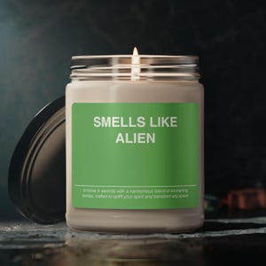 Può includere: Una candela bianca in un barattolo di vetro con un'etichetta verde su cui è scritto "SMELLS LIKE ALIEN". L'etichetta include anche il testo "Immerse in serenity with a harmonious blend of enchanting aromas, crafted to uplift your spirit and transform any space."