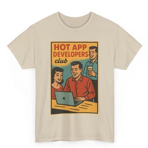 May include: Beige t-shirt featuring a retro-style graphic. The graphic reads "HOT APP DEVELOPERS club" and depicts a man using a laptop with a woman and another man holding a drink. The design has a vintage, comic book aesthetic.