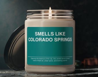 Op de afbeelding: Een glazen potkaars met een teal label waarop staat "Smells Like Colorado Springs". Het label vermeldt ook "Capturing the essence of the city, this candle blends vibrant notes of fresh air, urban spice, and evening warmth."