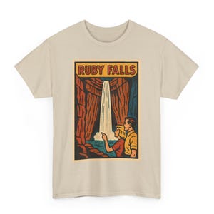 Op de afbeelding: Beige T-shirt met een retro graphic van Ruby Falls. Het ontwerp toont een waterval, een stel en de tekst "RUBY FALLS" in een rechthoekige lijst. Het kunstwerk gebruikt een vintage kleurenpalet van rood, blauw en geel.