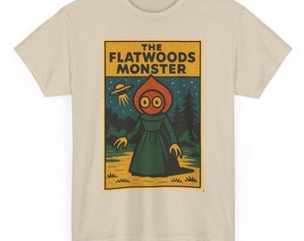 May include: Beige t-shirt featuring a graphic of the Flatwoods Monster. The design includes the text "THE FLATWOODS MONSTER" above an illustration of the creature, a UFO, and a forest scene. The monster has a red head and green dress.