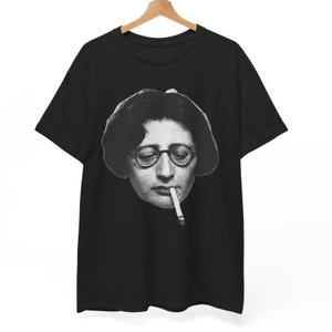 Può includere: T-shirt nero con immagine in bianco e nero di una persona con occhiali e sigaretta. L'immagine è un primo piano del volto della persona. La maglietta è appesa a una gruccia di legno.