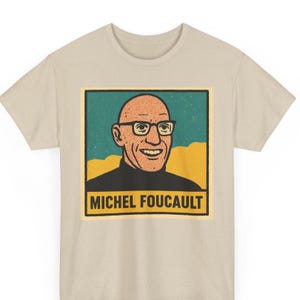 May include: Beige t-shirt featuring a retro-style portrait of Michel Foucault. The portrait is in a square frame with a teal and yellow background. The text "MICHEL FOUCAULT" is printed in a yellow rectangle below the portrait.