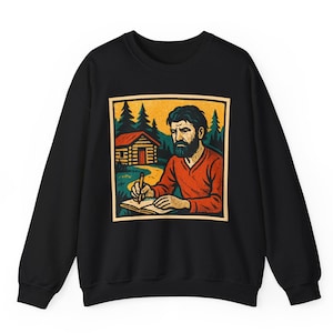 May include: Black sweatshirt featuring a framed illustration of a bearded person writing in a notebook. The illustration includes a cabin, trees, and a yellow and orange sky. The person is wearing a red shirt.