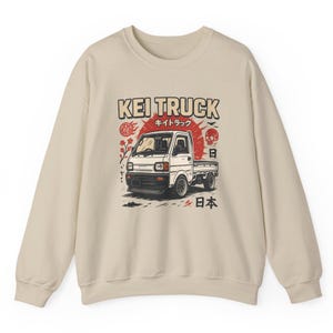 Pu&ograve; includere: Felpa beige con una grafica di un camion Kei bianco. Il design include il testo "KEI TRUCK" in marrone, caratteri giapponesi, un sole rosso, un teschio e simboli giapponesi.