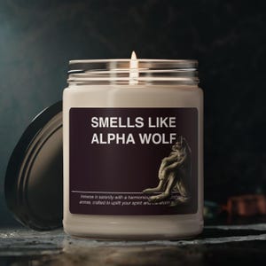 May include: A glass candle jar with a black lid. The candle is white and has a label that says "Smells Like Alpha Wolf". The label also features a wolf illustration.