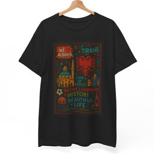 Może przedstawiać: Czarny t-shirt z kolorowym wzorem graficznym. Projekt zawiera tekst "We Love Albania" i "Tirane", a także orła i inne albańskie symbole. Koszulka wisi na drewnianym wieszaku.