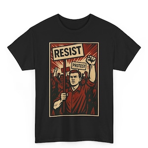 May include: Black t-shirt featuring a vintage-style graphic of a protest scene. The design includes a crowd of people, a person holding a sign that says "RESIST" and "PROTEST", and a raised fist. The artwork is in shades of red, beige, and black.
