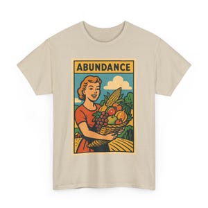 May include: Beige t-shirt featuring a retro-style graphic. The graphic depicts a woman holding a basket overflowing with fruits and vegetables, with the word "ABUNDANCE" above. The design is in a vintage comic book style.