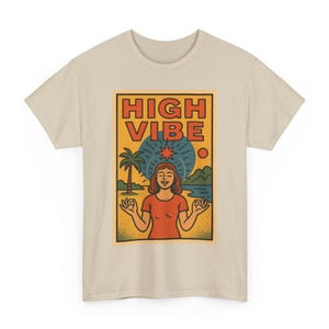 May include: Beige t-shirt featuring a graphic with the words "HIGH VIBE" in orange. The graphic depicts a woman meditating with a beach scene, including a palm tree, sun, and water. The shirt is made of a soft, light-colored material.