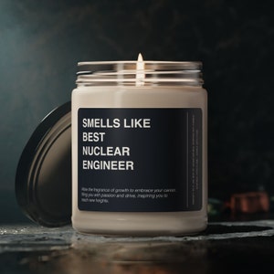 Pode incluir: Uma vela acesa, de cor creme, num frasco de vidro com tampa prateada. O rótulo diz "SMELLS LIKE BEST NUCLEAR ENGINEER". A vela está sobre uma superfície escura. O texto abaixo diz "Allow the fragrance of growth to embrace your career..."