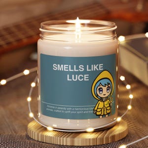 Könnte beinhalten: Eine brennende Kerze in einem Glas mit einem blauen Etikett mit der Aufschrift "SMELLS LIKE LUCE". Das Etikett zeigt eine Cartoonfigur in einem gelben Regenmantel. Die Kerze steht auf einem Holzuntersetzer, umgeben von Lichterketten.