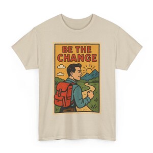 May include: Beige t-shirt with a retro graphic. The graphic features a person with a red backpack walking on a path towards mountains and a rising sun. The text "BE THE CHANGE" is at the top of the graphic.