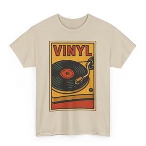 Peut inclure: T-shirt beige avec un motif rétro d'un tourne-disque vinyle. Le motif est dans un cadre rectangulaire avec le mot "VINYL" en haut. Le tourne-disque est noir et jaune avec un centre rouge.