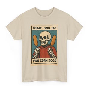 May include: Beige t-shirt featuring a graphic of a skeleton holding two corn dogs. The skeleton is wearing a red shirt. Text on the graphic reads "TODAY I WILL EAT TWO CORN DOGS."