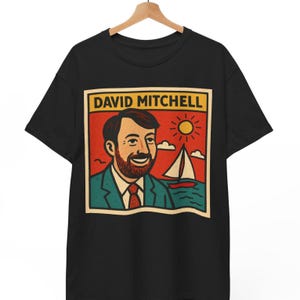 May include: Black t-shirt featuring a retro-style graphic of a man with a beard, a sailboat, and a sun. The graphic is framed by a yellow border with the text "DAVID MITCHELL" at the top. The man is wearing a suit and tie.