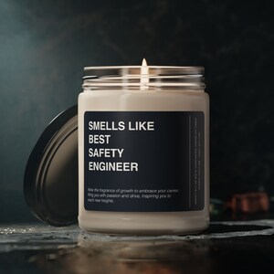 Pode incluir: Uma vela branca num frasco de vidro com um rótulo preto que diz "Smells Like Best Safety Engineer". O rótulo também inclui o texto "Allow the fragrance of growth to embrace your career, filling you with passion and drive, inspiring you to reach new heights."