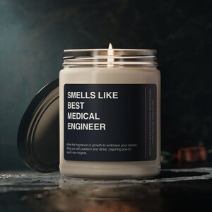 Pode incluir: Uma vela branca acesa num frasco de vidro com um rótulo preto. O rótulo diz "SMELLS LIKE BEST MEDICAL ENGINEER". O texto abaixo diz "Allow the fragrance of growth to embrace your career, filling you with passion and drive, inspiring you to reach new heights."