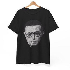 May include: Black t-shirt featuring a black and white portrait of a man wearing glasses and smoking a cigarette. The image is centered on the shirt, with the t-shirt hanging on a wooden hanger against a white background.