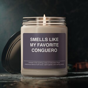 Puede incluir: Una vela blanca en un tarro de vidrio con una etiqueta negra que dice "Smells Like My Favorite Conguero". La etiqueta también incluye el texto "Immerse in the soothing notes of Instrument Player. A harmonious blend of soft wood, warm spices, and calming melody."