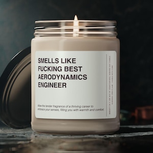 Puede incluir: Un tarro de vela de vidrio marrón con una etiqueta blanca que dice "Smells Like Fucking Best Aerodynamics Engineer". La vela está encendida y la cera se está derritiendo.