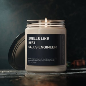 Pode incluir: Uma vela acesa num frasco de vidro com um rótulo preto que diz "SMELLS LIKE BEST SALES ENGINEER". O frasco está aberto, com a tampa ao lado. O texto abaixo diz "Allow the fragrance of growth to embrace your career."