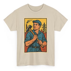 May include: Beige t-shirt featuring a retro-style illustration of a worker holding a sledgehammer. The artwork, set within a rectangular frame, depicts a man in blue work clothes, a cap, and industrial elements like smokestacks and power lines.