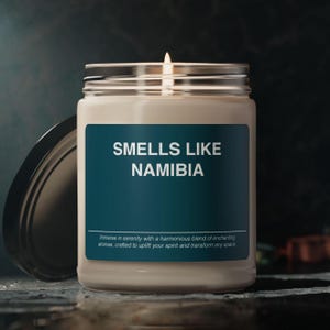 Puede incluir: Una vela en un tarro de vidrio con una tapa negra. La etiqueta en el tarro dice "Smells Like Namibia" e incluye el texto "Immerse in serenity with a harmonious blend of enchanting aromas, crafted to uplift your spirit and transform any space."