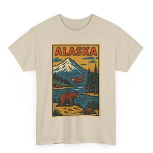 May include: A beige t-shirt featuring a rectangular graphic with the word "ALASKA" in red letters. The graphic depicts a mountain, lake, bear, and a red airplane. The design has a vintage, travel-poster style.