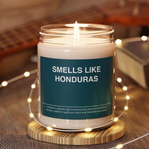 May include: A lit candle in a glass jar with a teal label that reads "SMELLS LIKE HONDURAS". The candle sits on a wooden coaster and is surrounded by string lights. The text "Immerse in serenity..." is printed on the label.