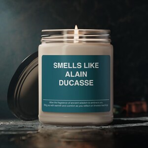 Op de afbeelding: Een witte kaars met een teal label waarop "Smells Like Alain Ducasse" staat. Het label bevat ook de tekst "Allow the fragrance of ancient wisdom to embrace you, filling you with warmth and comfort as you reflect on timeless teachings."
