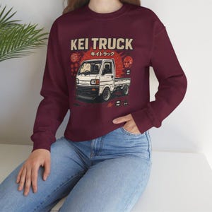 Pu&ograve; includere: Felpa girocollo bordeaux con grafica di un camion Kei bianco e testo giapponese. Il design include un sole rosso e dettagli a teschio. Il testo "KEI TRUCK" &egrave; visualizzato sopra l'illustrazione del camion.