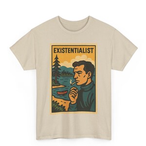 May include: Beige t-shirt featuring a rectangular graphic with the word "EXISTENTIALIST" above an illustration of a man smoking a cigarette by a lake. The man is wearing a blue sweater, and the background includes trees, water, and a small boat.