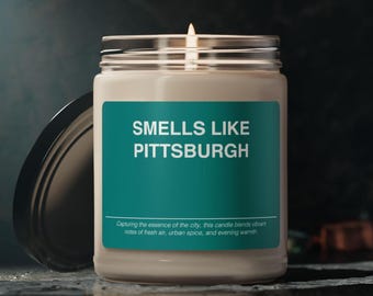 May include: A white candle in a glass jar with a teal label that reads "Smells Like Pittsburgh". The label also includes the text "Capturing the essence of the city, this candle blends vibrant notes of fresh air, urban spice, and evening warmth."
