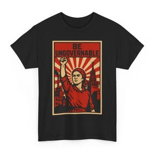 May include: Black t-shirt featuring a red and beige graphic. The graphic depicts a woman with a raised fist, the text "BE UNGOVERNABLE", and a background of buildings and sun rays. The shirt is made of a soft material.