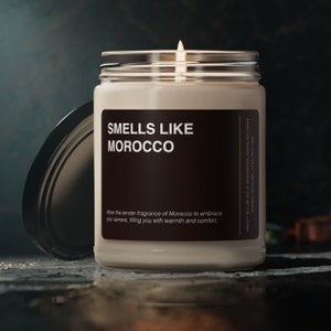 Może przedstawiać: Szklana świeca w słoiku z brązową etykietą, na której widnieje napis "SMELLS LIKE MOROCCO". Etykieta zawiera również tekst "Allow the tender fragrance of Morocco to embrace your senses, filling you with warmth and comfort." Świeca jest zapalona i płomień jest widoczny.