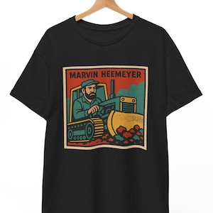 May include: Black t-shirt with a retro-style illustration of a man driving a bulldozer. The illustration is in a red, yellow, and blue color scheme and includes the text "Marvin Heemeyer".