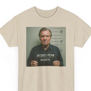Puede incluir: Una camiseta beige con una imagen de estilo foto policial de Jacques Pépin. La imagen lo muestra sosteniendo un cartel que dice "JACQUES PÉPIN 042875".