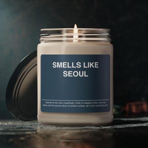 May include: A glass candle jar with a beige label that reads "SMELLS LIKE SEOUL". The label is navy blue with white text. The candle is lit and the jar has a black lid.