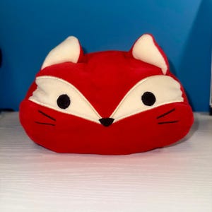 May include: A plush toy shaped like a fox head with bright red fabric, cream-colored accents around the eyes and ears, two black circular eyes, a small black triangular nose, and three stitched black whiskers on each side of the face.