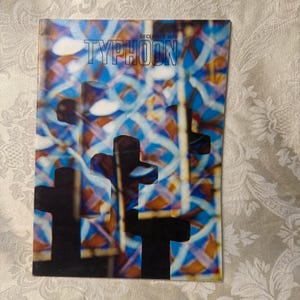 Könnte beinhalten: Ein Vintage-Magazin-Cover mit dem Titel "TYPHOON" in blauen Buchstaben. Der Hintergrund zeigt ein Buntglasfenster-Design mit blauen, orangefarbenen und weißen Farben. Silhouetten schwarzer Kreuze befinden sich im Vordergrund. Das Datum "DEZEMBER 1970" steht oben.
