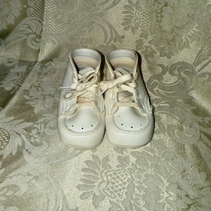 Puede incluir: Un par de zapatos de bebé color crema con cordones. Los zapatos tienen un diseño clásico con punta redondeada y pequeñas perforaciones. Los zapatos están hechos de un material suave y parecen ser vintage.