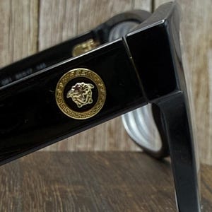 May include: Black sunglasses featuring a thick frame and a prominent gold-toned circular emblem on the temple. The emblem displays a detailed Medusa head motif surrounded by a Greek key pattern. The interior arm shows printed model information.